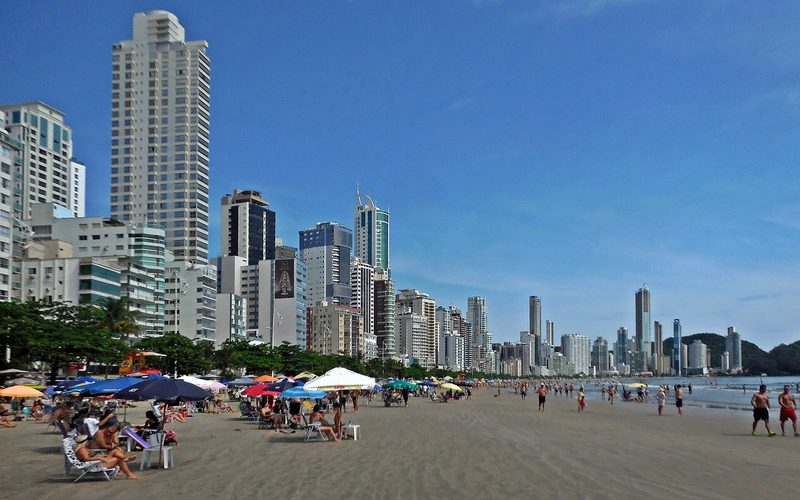 Praia com faixa de areia estreita e fileira de edifícios altos ao fundo, em Balneário Camboriú.