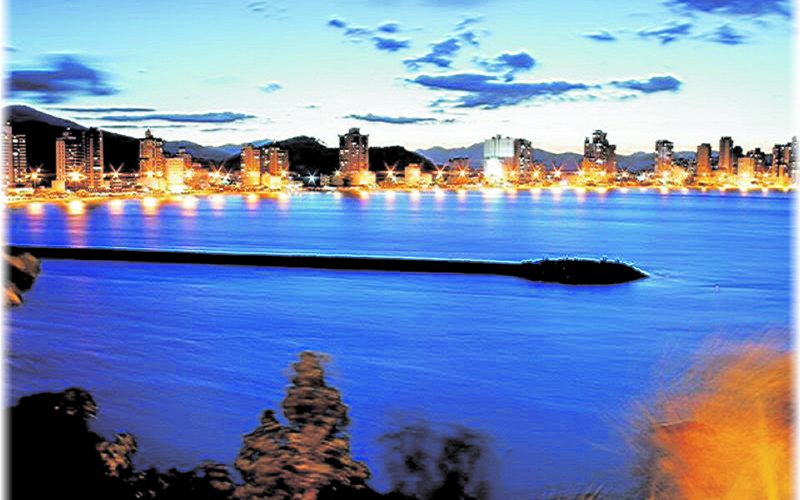 Faixa de praia iluminada à noite com muitos edifícios altos ao fundo.