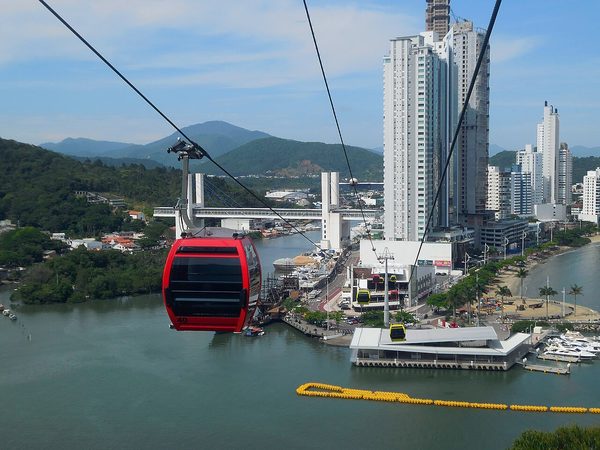 Cabine de teleférico sobre morro arborizado com vista para a praia de Balneário Camboriú.