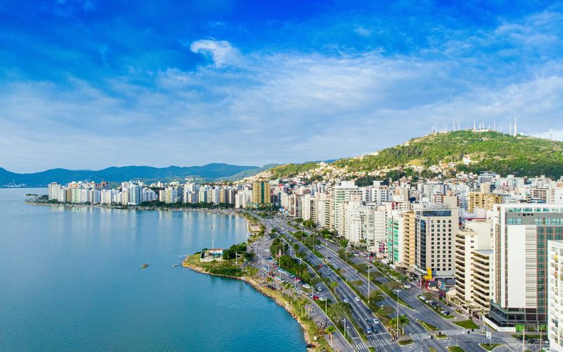 Vista aérea da orla da Beira-Mar Norte, em Florianópolis, com prédios altos entre a avenida e a baía.