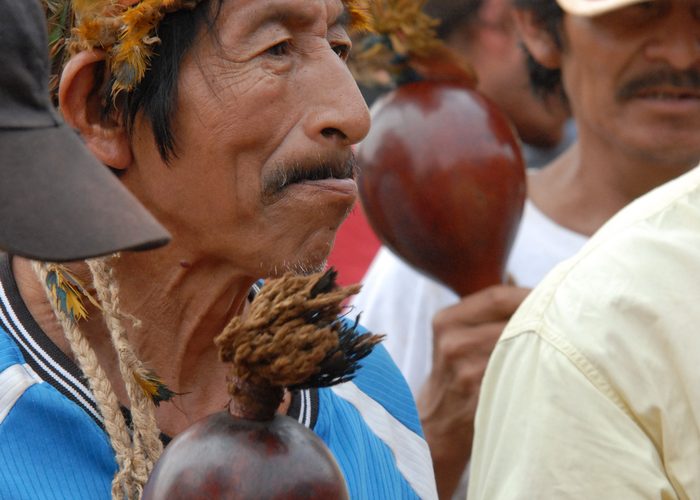 Homens guarani com cocares de penas tocando maracás durante uma celebração.