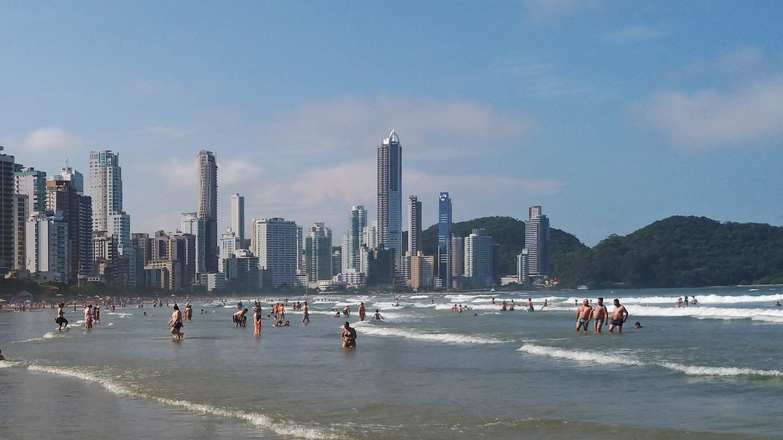 Praia de areia clara com mar aberto e morros cobertos de vegetação ao fundo, no litoral de Santa Catarina.